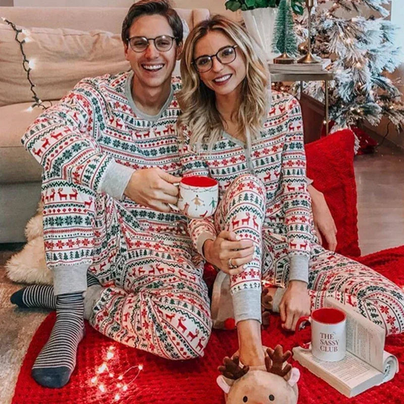 Couple wearing matching festive pajamas sitting on a red blanket with a Christmas tree in the background.